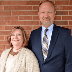 Two team members standing outside against a brick wall, smiling.